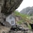 A small wooden house nestled under a massive rocky overhang. The house appears weathered with white-framed windows, surrounded by large boulders and rock formations. In the background, a lush green landscape and distant cliffs with a waterfall create a serene and secluded atmosphere.