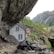 A small wooden house nestled under a massive rocky overhang. The house appears weathered with white-framed windows, surrounded by large boulders and rock formations. In the background, a lush green landscape and distant cliffs with a waterfall create a serene and secluded atmosphere.
