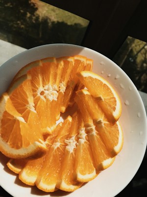 Slices of bright orange fruit arranged neatly in a white bowl. The sunlight casts a warm glow over the fruits, highlighting their juicy texture and vibrant color. The backdrop includes a dimly lit window with a hint of greenery outside.