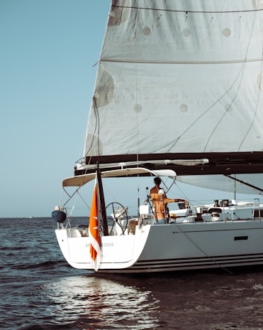 A sailing yacht with large sails is navigating in the open sea. Two people are visible on the deck, engaged in operating the vessel. The back of the yacht displays an orange flag, and the waters around the boat are calm under a clear blue sky.