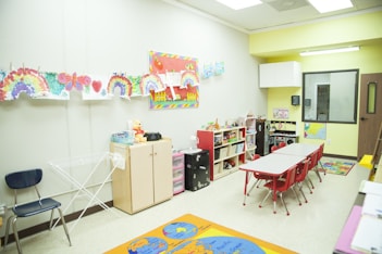 The image depicts a colorful classroom setting with a row of tables and red chairs. Various artworks, including rainbows and hearts, are displayed on the wall. Shelves and cabinets contain toys and games, and there are educational materials like maps and charts. A cheerful, inviting atmosphere is created by the bright decorations and organized layout.