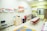 The image depicts a colorful classroom setting with a row of tables and red chairs. Various artworks, including rainbows and hearts, are displayed on the wall. Shelves and cabinets contain toys and games, and there are educational materials like maps and charts. A cheerful, inviting atmosphere is created by the bright decorations and organized layout.