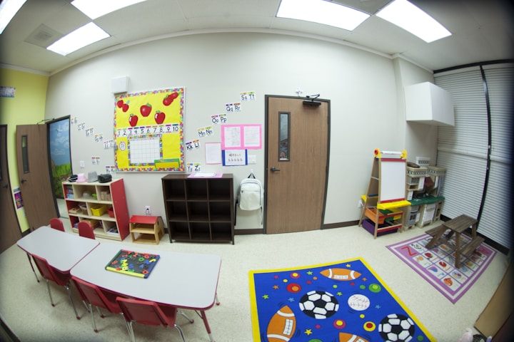 A bright, organized classroom with educational materials on the walls. There are colorful alphabet and number charts, a variety of shelves holding different toys, a small table with red chairs, an easel, and a vibrant blue rug featuring sports balls. The room has a welcoming and educational atmosphere.