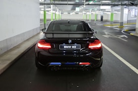 A black BMW M2 car is parked in an indoor parking garage. The car's rear license plate is visible with the text 'BMW M2'. The parking area has concrete walls, supporting pillars painted in green and white, and overhead fluorescent lighting.