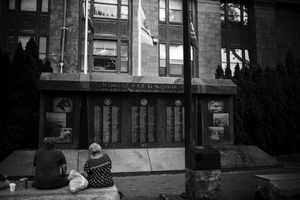 grayscale photo of man and woman sitting on bench near building