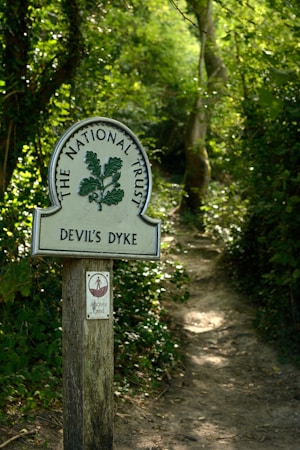 A wooden post with a sign indicates a pathway leading through a lush, green wooded area. The sign reads 'The National Trust' and 'Devil's Dyke' with a small secondary sign indicating access land.