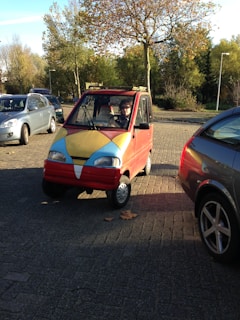 A small, colorful vehicle with a red body and yellow and blue accents is parked on a paved area surrounded by other cars. A person is sitting inside the vehicle, which is set against a backdrop of trees and a clear sky.