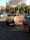 A small, colorful vehicle with a red body and yellow and blue accents is parked on a paved area surrounded by other cars. A person is sitting inside the vehicle, which is set against a backdrop of trees and a clear sky.