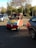 A small, colorful vehicle with a red body and yellow and blue accents is parked on a paved area surrounded by other cars. A person is sitting inside the vehicle, which is set against a backdrop of trees and a clear sky.