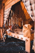A rustic wooden cabin interior featuring a simple bed covered with fur throws and woolen blankets. The bed is supported by a basic wooden frame, and curtain partitions made from checkered fabric hang on either side. The walls are constructed from vertical wooden logs, giving off a cozy, earthy atmosphere.