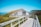 A wooden pathway leads to a rustic building with a thatched grass roof, set in a scenic landscape. Surrounded by grass and wildflowers, the structure has a traditional, earthy design with wooden panels. In the background, gentle hills and a clear blue sky expand the tranquil view.