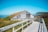 A wooden pathway leads to a rustic building with a thatched grass roof, set in a scenic landscape. Surrounded by grass and wildflowers, the structure has a traditional, earthy design with wooden panels. In the background, gentle hills and a clear blue sky expand the tranquil view.