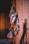 A rustic wooden interior features several hanging sacks, bundles of cloth, and what appears to be an animal fur. The wooden walls and floor create a warm, natural ambiance, enhanced by lighting that casts soft shadows. In the background, there are wooden shelves with pottery or other household items.