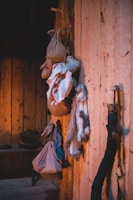 A rustic wooden interior features several hanging sacks, bundles of cloth, and what appears to be an animal fur. The wooden walls and floor create a warm, natural ambiance, enhanced by lighting that casts soft shadows. In the background, there are wooden shelves with pottery or other household items.