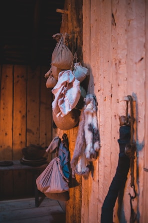 A rustic wooden interior features several hanging sacks, bundles of cloth, and what appears to be an animal fur. The wooden walls and floor create a warm, natural ambiance, enhanced by lighting that casts soft shadows. In the background, there are wooden shelves with pottery or other household items.