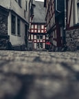 A narrow cobblestone alleyway flanked by half-timbered houses leads to a charming, traditional building at the end. The architecture showcases a combination of exposed wood beams and white plaster, creating a classic aesthetic. The atmosphere is calm and nostalgic, with muted colors and overcast lighting adding to the historic charm.