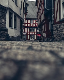 A narrow cobblestone alleyway flanked by half-timbered houses leads to a charming, traditional building at the end. The architecture showcases a combination of exposed wood beams and white plaster, creating a classic aesthetic. The atmosphere is calm and nostalgic, with muted colors and overcast lighting adding to the historic charm.