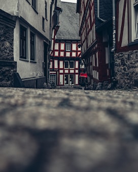 A narrow cobblestone alleyway flanked by half-timbered houses leads to a charming, traditional building at the end. The architecture showcases a combination of exposed wood beams and white plaster, creating a classic aesthetic. The atmosphere is calm and nostalgic, with muted colors and overcast lighting adding to the historic charm.