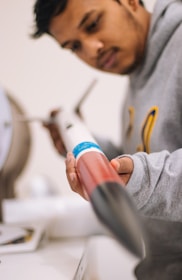 A person is examining a model rocket, holding it carefully in their hand. The rocket is predominantly red and white with a blue band around its body. The figure is wearing a gray hoodie and appears focused on the rocket.