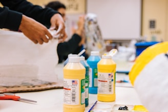 A worktable is set up for an art project, including bottles of acrylic paint in various colors like yellow and blue, paintbrushes, and palettes. A person is engaged in a crafting activity, using materials such as paper or fabric. There are other art supplies visible on the table.