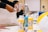 A worktable is set up for an art project, including bottles of acrylic paint in various colors like yellow and blue, paintbrushes, and palettes. A person is engaged in a crafting activity, using materials such as paper or fabric. There are other art supplies visible on the table.