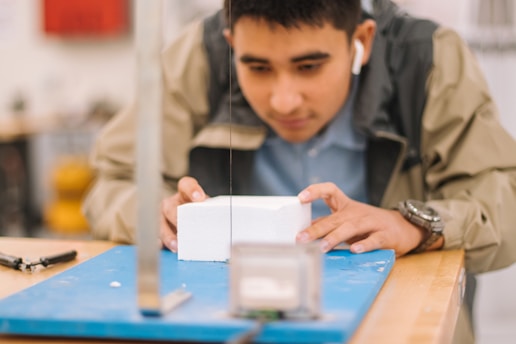 A person is carefully observing and handling a white block placed on a blue surface in what appears to be a workshop or lab environment. The individual is wearing earbuds and a jacket, focusing intently on the task at hand.
