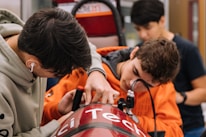 Three individuals are working together on a technical project, focused on adjusting a red device with the word 'Tech' partially visible. They appear to be in a workshop or lab environment, with various tools and equipment visible in the background. The person on the left is wearing a hoodie and earphones, while the person in the middle is in an orange hoodie, intently working on the project.