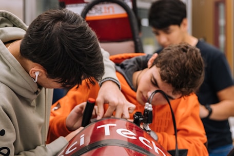 Three individuals are working together on a technical project, focused on adjusting a red device with the word 'Tech' partially visible. They appear to be in a workshop or lab environment, with various tools and equipment visible in the background. The person on the left is wearing a hoodie and earphones, while the person in the middle is in an orange hoodie, intently working on the project.