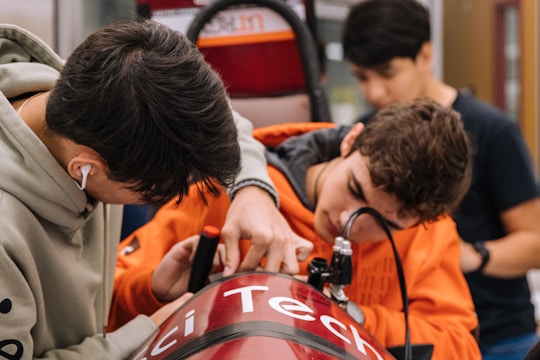 Three individuals are working together on a technical project, focused on adjusting a red device with the word 'Tech' partially visible. They appear to be in a workshop or lab environment, with various tools and equipment visible in the background. The person on the left is wearing a hoodie and earphones, while the person in the middle is in an orange hoodie, intently working on the project.