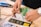 Hands are working on a breadboard circuit with numerous green wires looping over it. Various tools and wires are scattered around the worktable, including a pair of wire cutters and a watch on the wrist of the person working.