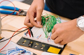 Hands are working on a breadboard circuit with numerous green wires looping over it. Various tools and wires are scattered around the worktable, including a pair of wire cutters and a watch on the wrist of the person working.