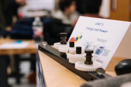 A classroom setting featuring a wooden table with a mechanical setup involving gears and rods. A paper titled 'UNIT 1 Energy and Power' is displayed, containing diagrams and text. In the background, blurred figures and classroom materials can be seen.