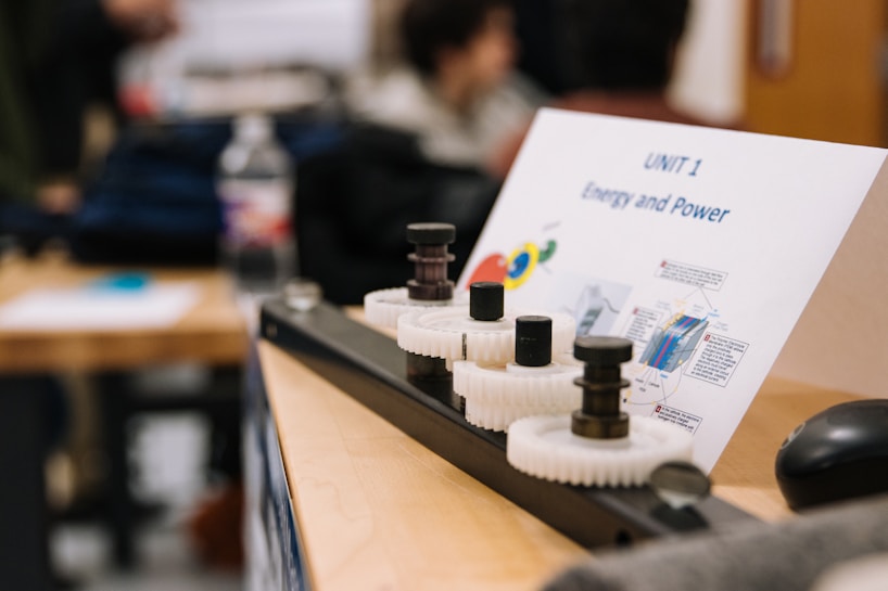 A classroom setting featuring a wooden table with a mechanical setup involving gears and rods. A paper titled 'UNIT 1 Energy and Power' is displayed, containing diagrams and text. In the background, blurred figures and classroom materials can be seen.
