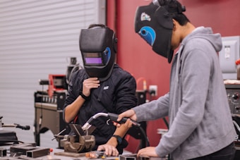 Two individuals wearing welding helmets are engaged in a metalworking activity in a workshop. One person holds a welding tool over a workbench, while the other observes. Various tools and equipment are scattered around the workspace, and the background shows metallic surfaces and workshop machinery.