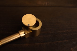A brass smoking pipe rests on a dark wooden surface. The pipe features a cylindrical bowl with a hinged lid partially open, revealing the mesh inside. The warm metallic color contrasts against the deep brown wooden background.