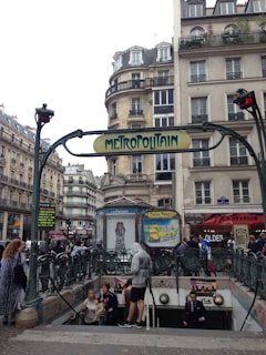 An urban scene featuring a stone building in Paris with wrought iron balconies and a classic 'Metropolitain' metro sign in Art Nouveau style. Several people are entering and exiting the metro station beneath the sign, while others are walking on the pavement. The background includes typical Parisian architecture with multiple stories and shops lining the street.