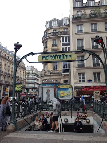 An urban scene featuring a stone building in Paris with wrought iron balconies and a classic 'Metropolitain' metro sign in Art Nouveau style. Several people are entering and exiting the metro station beneath the sign, while others are walking on the pavement. The background includes typical Parisian architecture with multiple stories and shops lining the street.