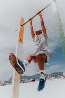 A person engages in physical activity by hanging from a pull-up bar. The scene takes place on a sunny beach with people and colorful umbrellas in the background. The person wears blue sneakers, socks with stripes, and patterned shorts.
