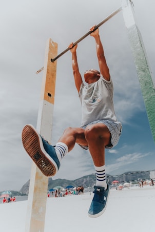 A person engages in physical activity by hanging from a pull-up bar. The scene takes place on a sunny beach with people and colorful umbrellas in the background. The person wears blue sneakers, socks with stripes, and patterned shorts.