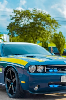 A blue sports car with black rims parked on a road, featuring yellow accents and the word 'POLICE' visible on the front. The car has a sleek design, with distinctive round headlights and a logo at the front. In the background, there are green trees and a clear blue sky.