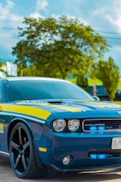 A blue sports car with black rims parked on a road, featuring yellow accents and the word 'POLICE' visible on the front. The car has a sleek design, with distinctive round headlights and a logo at the front. In the background, there are green trees and a clear blue sky.