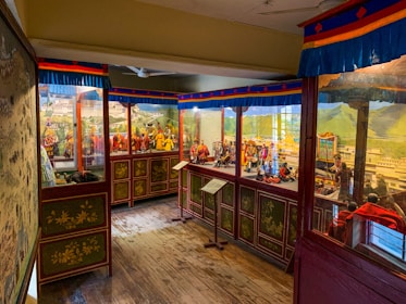 A room in a museum with glass cases displaying intricate dioramas portraying historical or cultural scenes. The walls are adorned with detailed murals, and there is wood paneling at the bottom. Bright colors, primarily blue, red, and yellow, accentuate the displays.
