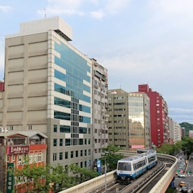 An urban scene featuring a train on an elevated track traveling alongside a line of multi-story buildings. The architecture includes a mix of modern glass windows and older-style structures. There are green trees lining the street below, and a mix of cars can be seen on the road. The sky is partly cloudy, adding a soft light to the scene.