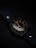 A watch with a dark leather strap and a metallic case, featuring a black dial with contrasting white and gold numerals and hands, is positioned at an angle. The background is subtly lit in dark tones, enhancing the sleek and elegant design of the watch.