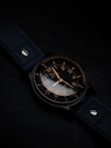 A watch with a dark leather strap and a metallic case, featuring a black dial with contrasting white and gold numerals and hands, is positioned at an angle. The background is subtly lit in dark tones, enhancing the sleek and elegant design of the watch.
