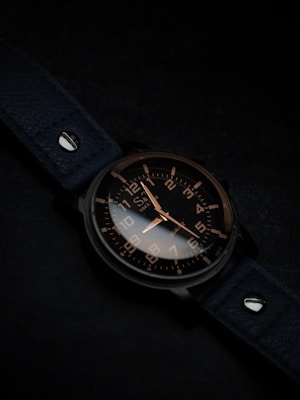 A watch with a dark leather strap and a metallic case, featuring a black dial with contrasting white and gold numerals and hands, is positioned at an angle. The background is subtly lit in dark tones, enhancing the sleek and elegant design of the watch.