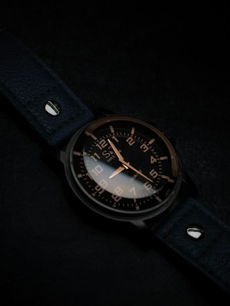 A watch with a dark leather strap and a metallic case, featuring a black dial with contrasting white and gold numerals and hands, is positioned at an angle. The background is subtly lit in dark tones, enhancing the sleek and elegant design of the watch.