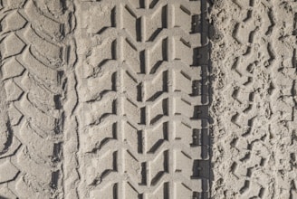 Three distinct tire tracks are imprinted in the sandy ground, each with a unique tread pattern. The left track has a wavy design, the center track features a more angular, interlocking pattern, and the right track shows irregular, more eroded patterns.