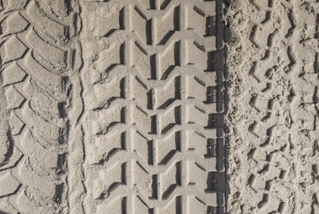 Three distinct tire tracks are imprinted in the sandy ground, each with a unique tread pattern. The left track has a wavy design, the center track features a more angular, interlocking pattern, and the right track shows irregular, more eroded patterns.