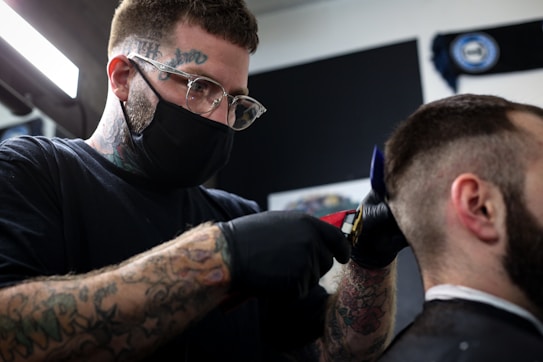 A barber with tattoos on his arms and forehead is wearing a mask and gloves while using clippers to cut a customer's hair. The setting appears to be a barber shop, with a dark background and bright lighting focused on the activity.